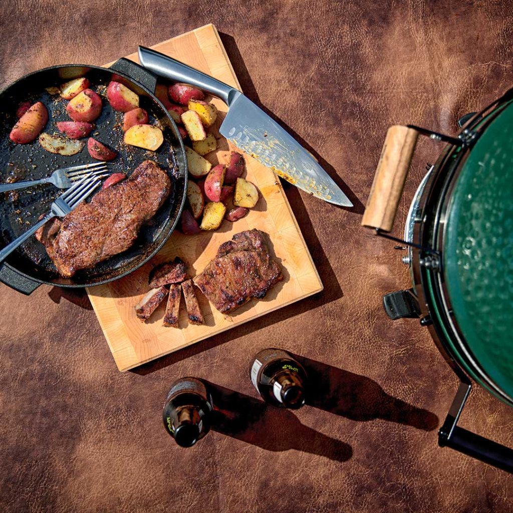 Cast Iron Skillet with steak, potatoes, forks and knife on cutting and Big Green Egg.